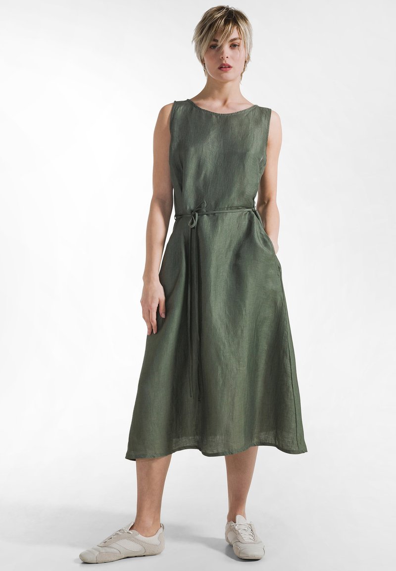 Vestido de lino verde oliva hasta la rodilla con escote redondo, cinturón de atar y bolsillos laterales. Combinado con zapatillas deportivas gris claro.