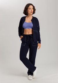 Marineblauw trainingspak met een ritsjack en een broek met trekkoord. De top is gecombineerd met een lichtpaarse sportbeha. Glad stof met contrasterende naden.