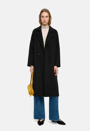 Kobieta ubrana w czarny długi płaszcz, beżową bluzkę na guziki, szerokie niebieskie dżinsy, czarne szpiczaste buty, trzymająca małą żółtą torebkę.