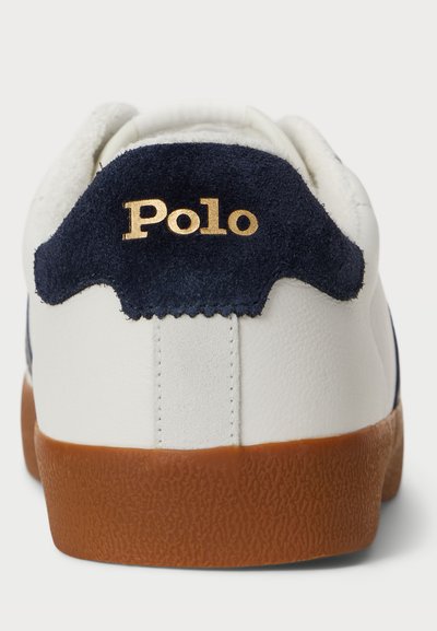 Valged tennisetooted, millel on tume sinine sametist konts, kuldne "Polo" logo ja tekstuuriga pruun kummist tald. Sile materjal ja klassikaline disain.