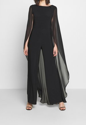 Woman wearing a black jumpsuit with wide legs and sheer cape-style sleeves, paired with black open-toe heels, standing on a gray floor.