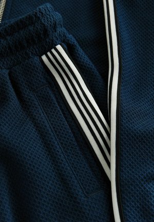 Marineblauer, strukturierter Sportbekleidungsstoff mit einem Netzmuster, der eine Reißverschlusstasche und schwarz-weiße Streifenakzente an der Seite aufweist.