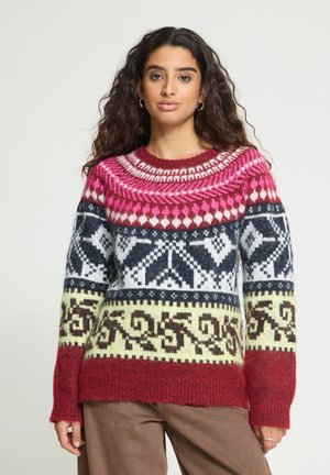 Femme aux longs cheveux bouclés porte un pull coloré à motifs géométriques rouges, roses, noirs, blancs et jaunes.