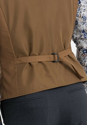 Gilet marron avec un dos ceinturé, en tissu texturé, associé à une chemise à motifs floraux dans des tons de bleu et de blanc.