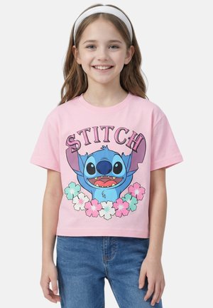 Fille souriante portant un bandeau blanc, un T-shirt rose avec le personnage Stitch et des motifs floraux, et un jean bleu, debout devant un fond uni.