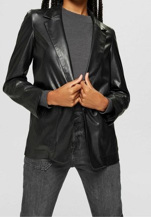 Persoon draagt een zwart imitatieleren blazer over een donkergrijze trui met ronde hals en zwarte jeans, houdt met beide handen de revers van het blazer vast.