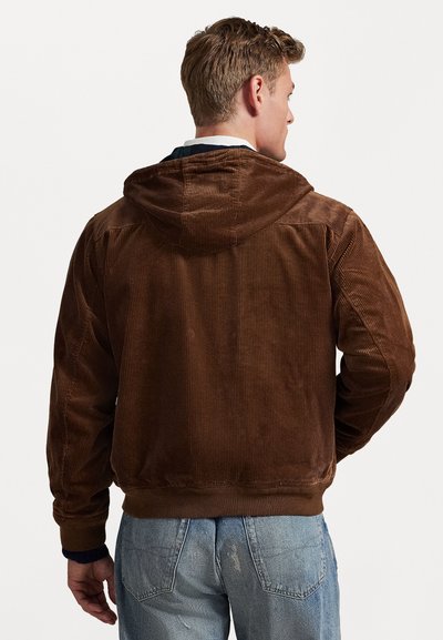 Brun kordfløjljakke med hætte, der har ribbet tekstur og elastiske manchetter, båret over lyseblå slidte jeans.