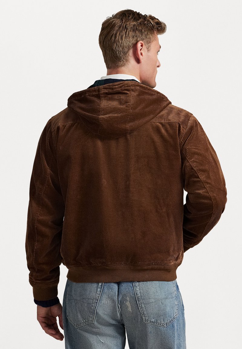 Giacca in corduroy marrone con cappuccio, caratterizzata da una texture a coste e polsini elasticizzati, indossata sopra jeans chiari strappati blu.