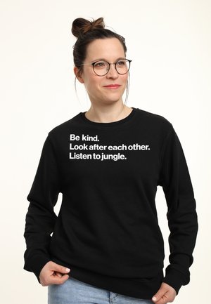 Sweatshirt noir avec du texte blanc : "Soyez gentils. Prenez soin les uns des autres. Écoutez la jungle." Matière en coton, col rond, manches longues.