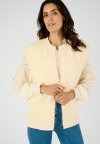 Chaleco sin mangas de color crema claro con un acabado suave y texturizado. Presenta un escote redondo y un cierre de botones, usado sobre una blusa estampada.