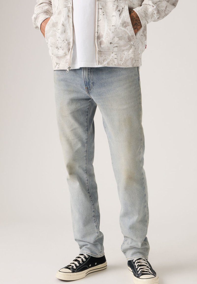 Φωτεινό γαλάζιο τζιν παντελόνι με ξεθωριασμένο φινίρισμα, σε συνδυασμό με μαύρα sneakers χαμηλού τύπου με λευκές σόλες. Το άτομο φοράει γκρι ζακέτα με φερμουάρ και μοτίβο.