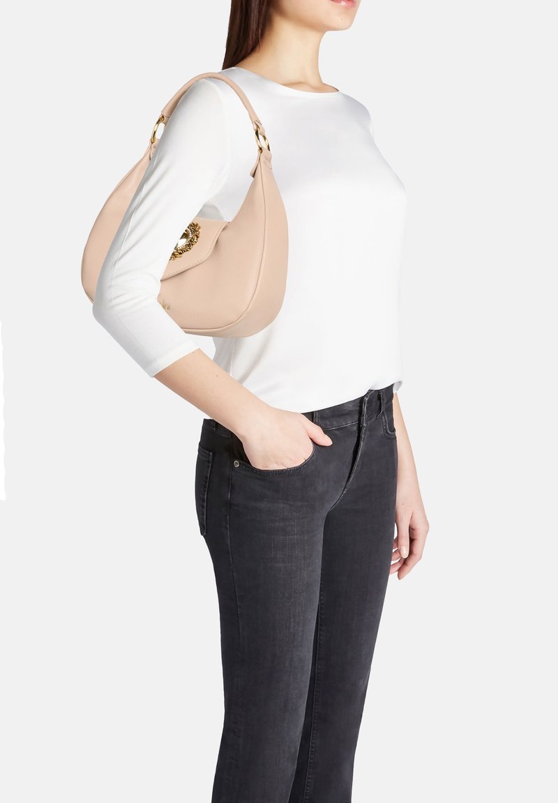 Borsa a spalla in pelle color tan con una forma curva, dettagli in metallo dorato e una chiusura decorativa. Il modello indossa un top bianco e jeans neri.
