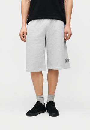 Mann trägt hellgraue knielange Shorts, schwarze Turnschuhe, graue Socken und ein schwarzes T-Shirt, steht vor einem schlichten hellen Hintergrund.