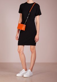 Svart klänning med kort ärm och figursydd design, parat med vita sneakers och en liten orange crossbody-väska med metalliska detaljer.