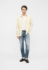 Chemise boutonnée jaune pâle portée sur un t-shirt blanc, associée à un jean bleu délavé et des mocassins foncés. Texture lisse, coupe décontractée.
