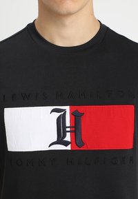 Czarna koszulka z dużą naszywką z haftowanym logo w kolorach czerwonym, białym i niebieskim, z napisem "LEWIS HAMILTON" oraz "TOMMY HILFIGER" poniżej.