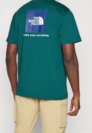 Osoba nosząca zielony t-shirt z logo "The North Face" i napisem "Never Stop Exploring" na plecach, w połączeniu z beżowymi szortami cargo.