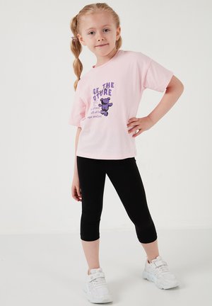 Noor neiu heledate punutud juustega poseerimas roosa T-särgi, kus on lilla tekst ja koala graafika, mustade retuuside ning valgete tossudega.