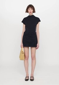 Sort turtleneck i svart, mørk mønstret miniskjørt og svarte ballerinas. Modellen holder en vevd gul håndveske. Enkel bakgrunn.