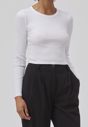Femme portant un crop top blanc ajusté à manches longues, rentré dans un pantalon noir ample taille haute avec fermeture à boutons.