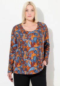 Langärmliges Oberteil mit Paisleymuster in orange, blau und braun; runder Ausschnitt und lässige Passform. Kombiniert mit schwarzen Hosen.