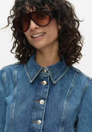 Kvinde med krøllet hår iført oversized mørke solbriller og en knappet denimjakke, der smiler mod en ensfarvet baggrund.