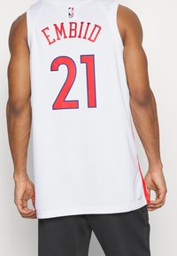 Maglia NBA bianca con accenti rossi. Sul retro è stampato "EMBIID" in rosso e "21" in grassetto rosso contornato di blu. Design senza maniche, tessuto leggero.