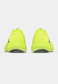 Ζευγάρι από νέον κίτρινα παπούτσια τρεξίματος, ορατά από πίσω, που δείχνουν παχιά σόλα και μαύρο σχέδιο πέλματος πάνω σε λευκό φόντο.