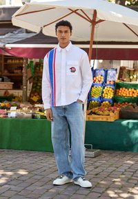 Hvid langærmet skjorte med blå og røde striber, frontlomme og patch; parret med lyseblå jeans og hvide sneakers på en brostensbelagt gade.