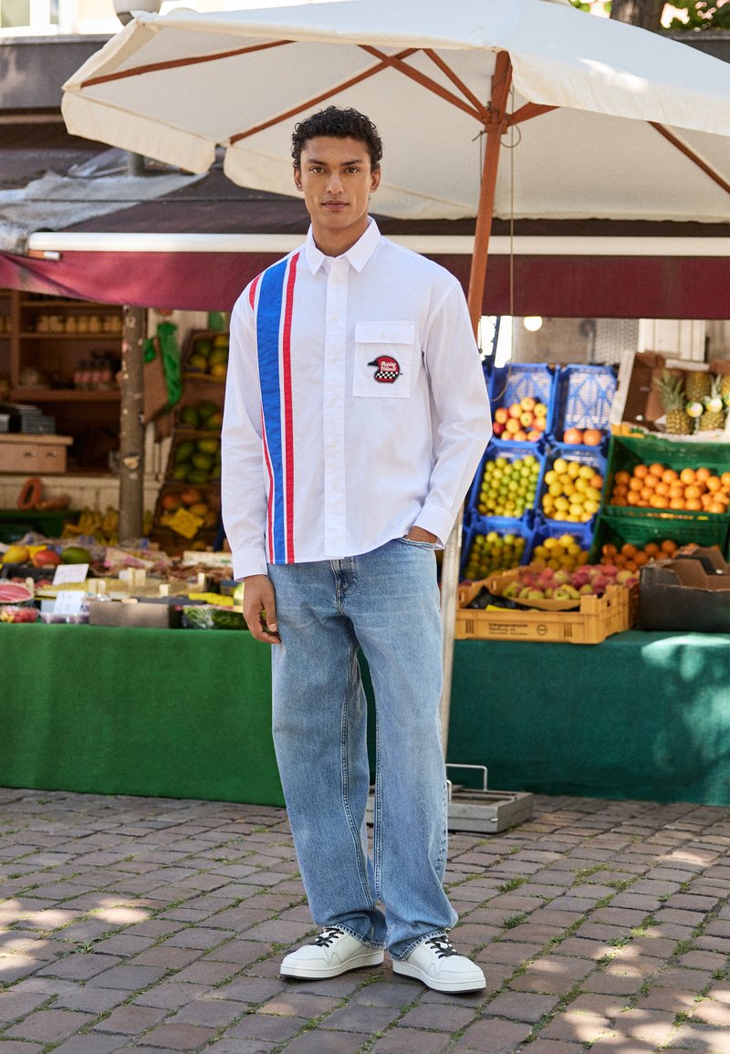 Hvid langærmet skjorte med blå og røde striber, frontlomme og patch; parret med lyseblå jeans og hvide sneakers på en brostensbelagt gade.