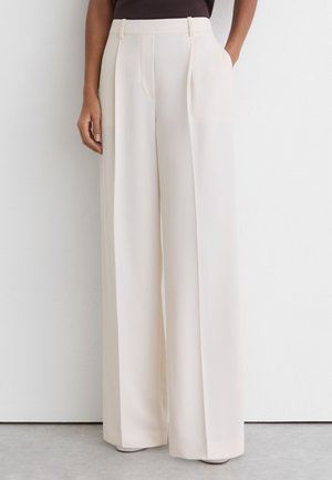 Mujer con pantalones cremosos de cintura alta y pata ancha con bolsillos laterales, combinados con una parte superior negra sobre un fondo blanco liso.