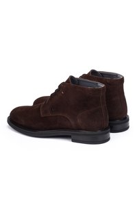 Un paio di stivali alla caviglia in suede marrone scuro con lacci neri e suole in gomma nera, mostrati da un angolo laterale e posteriore su sfondo bianco.