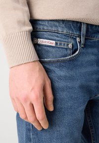 Bež pleteni pulover ima rukav preko plavih traperica, s crveno-bijelim Calvin Klein oznakom blizu džepa. Ruka je naslonjena na traperice.