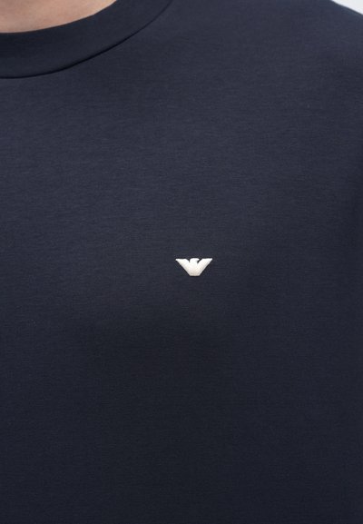 Tmavomodré tričko s krátkym rukávom z mäkkej elastickej látky, s malým bielym vyšívaným logom na hrudi. Klasický okrúhly výstrih.