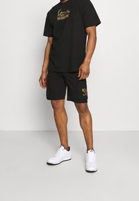 Camiseta de algodón negra con logo dorado, combinada con pantalones cortos negros con bolsillos laterales y texto dorado. Se lleva con zapatillas blancas.