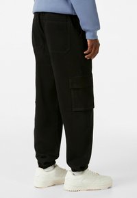 Pantaloni cargo neri con polsini elastici, dotati di tasche laterali e una finitura testurizzata. Indossati con sneakers bianche e una felpa azzurra chiara.