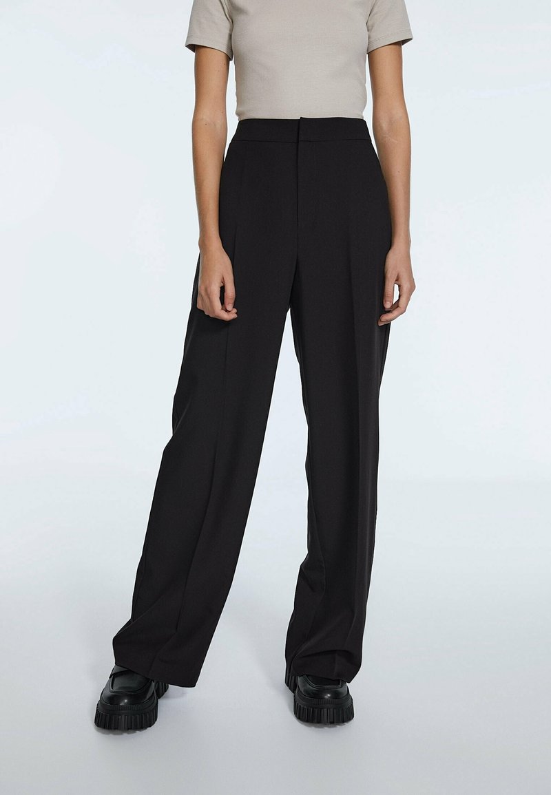 Personne portant un pantalon noir taille haute à jambes larges, un haut beige à manches courtes et des chaussures noires épaisses, debout devant un fond clair uni.