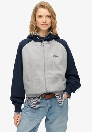 Gray and navy hoodie with zip closure, drawstring hood, and ribbed cuffs. Features a front logo and front pockets. Worn over a striped shirt.