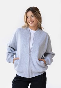 Szara bluza zapinana na zamek o luźnym kroju, z prążkowanymi mankietami i bocznymi kieszeniami, noszona na białej koszulce, na jednolitym tle.