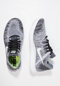 Scarpe sportive lavorate a maglia in bianco e nero con lacci neri, logo Swoosh bianco e sole verde all'interno di una suola bianca. Parte superiore traspirante e testurizzata.