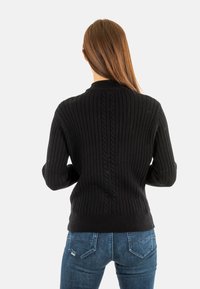 Pull en maille côtelée noir avec un col haut, présentant des détails en torsades le long de la colonne vertébrale. Assorti à un jean bleu, affichant une coupe ajustée.