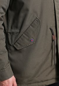 Veste vert olive avec une texture en surface, dotée d'une poche à patte diagonale et d'un accent logo circulaire. Comprend un détail à cordon.