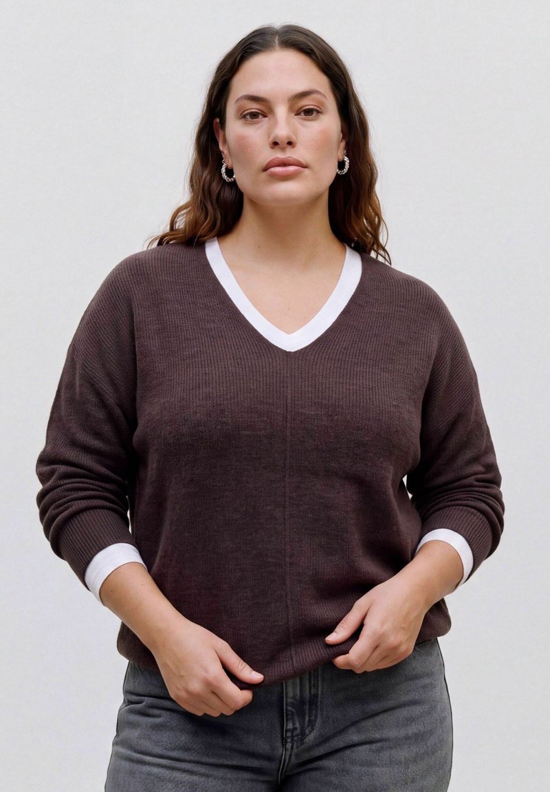 Femme aux longs cheveux ondulés portant un pull marron à col en V avec des bordures blanches et un jean gris, debout devant un fond clair uni.