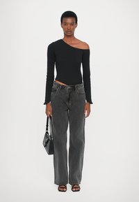 Top lungo asimmetrico nero a maniche lunghe abbinato a jeans larghi grigi scuri a vita alta. La modella tiene in mano una piccola borsa nera. Sandali neri piatti.