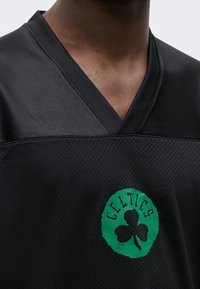 Εικόνα κοντινής λήψης ενός ατόμου που φοράει μαύρη αθλητική φανέλα με το πράσινο λογότυπο των Boston Celtics, που απεικονίζει ένα τριφύλλι στο στήθος.