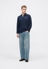 Jūnniecības garām piedurknēm polo krekls ar baltu apkakli, kas rotāts ar logotipu. Apvienots ar gaiši ziliem platām kājām džinsiem un melnām kurpēm. Vienkāršs, ikdienišķs dizains.