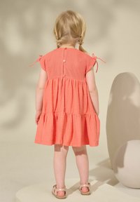 Korallefarbendes Baumwollkleid mit kurzen Cap-Ärmeln, einem gestuften Rock und einer Knopfleiste hinten. Geflochtenes Haar und Sandalen runden den Look ab.
