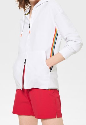 Weißer Reißverschluss-Hoodie mit Regenbogenstreifen entlang des Ärmels, vorderen Taschen und einer Kapuze mit Kordelzug; kombiniert mit roten Shorts.