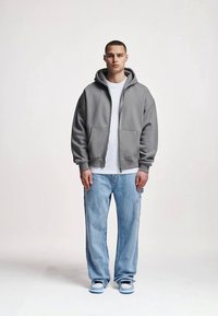 Pilka megztinė su užtrauktuku, po ja baltas marškinėlis, šviesiai mėlynos platėjančios džinsinės kelnės ir mėlynos bateliai. Modelis stovi rankomis palei šonus prieš vienodą foną.