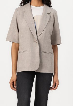 Femme portant un blazer beige à manches courtes sur un t-shirt blanc et un pantalon noir, montrée du cou jusqu'aux hanches sur un fond uni.